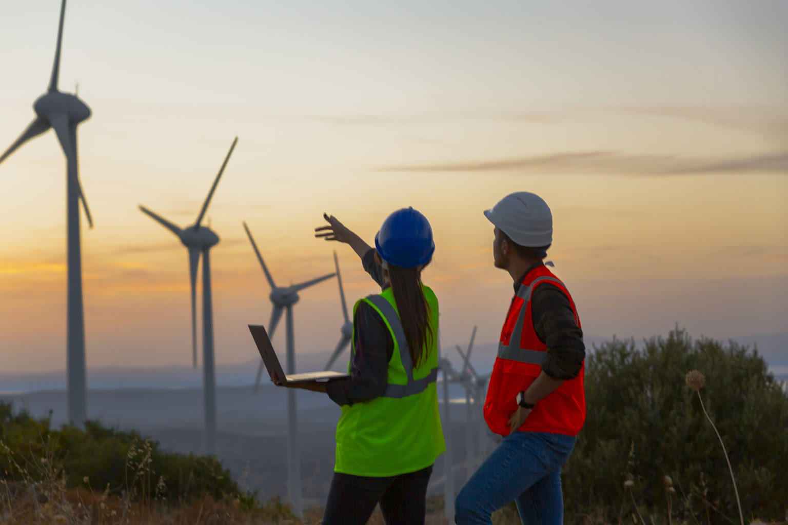 Young electrical engineer woman and business man standing in front of wind turbines checking and working about technical problems and writes the results of measurements with laptop pc in wind power plant electric energy station.